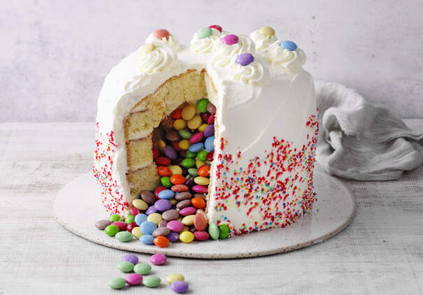Eine runde Piñata‑Torte mit weißer Creme ist mit bunten Schokolinsen gefüllt und mit Streuseln verziert.