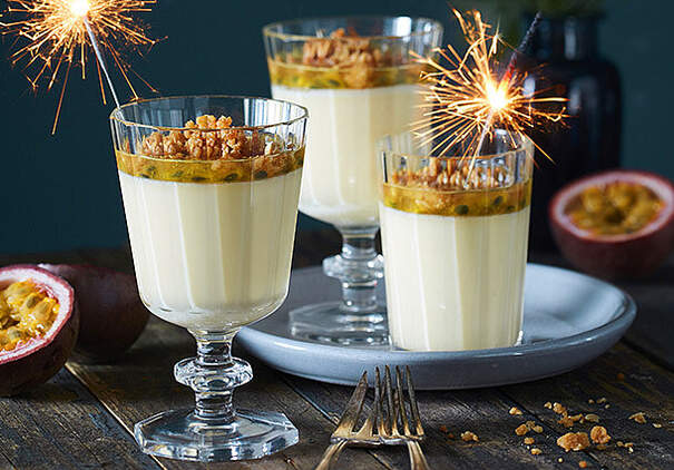 Zwei hohe Dessertgläser sind mit heller Panna Cotta gefüllt, darauf liegt eine gelbe Passionsfrucht‑Maracuja‑Schicht; kleine Wunderkerzen brennen darin
