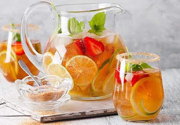 Auf einem hellgrauen Untergrund stehen ein großer Glaskrug und ein hohes Glas mit goldfarbenem Zitronen‑Limetten‑Eistee, der mit Eiswürfeln gefüllt ist.