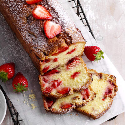 Ein länglicher Kuchen mit brauner und heller Marmorierung liegt angeschnitten auf einem Brett; darüber ist Puderzucker gestäubt und einige frische Erdbeeren zieren den Rand. Alt-Tag: Marmorkuchen mit Puderzucker und Erdbeeren
