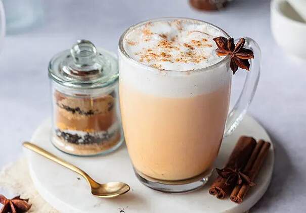 Auf dem Bild steht ein großes Glas Chai Latte auf einem kleinen weißen Kaffeeteller mit einem goldenen Löffel. An den Rand des Glases wurde dekorativ eine Zimtblüte gesteckt. Daneben steht ein kleines Glas mit einer Nachspeise (vermutlich Tiramisu) und auf dem Teller liegen Zimtstangen. 