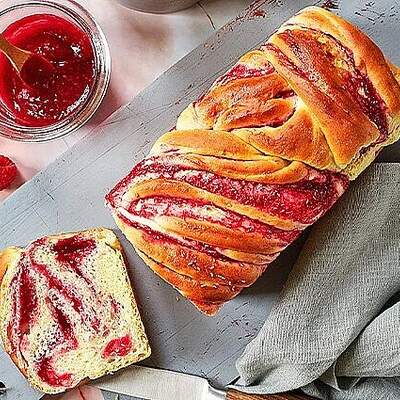 Auf einer Steinplatte liegt ein geflochtener Zopf aus Hefeteig mit rosa-weißem Himbeermuster; daneben stehen Schälchen mit Himbeersauce und eine Teekanne.