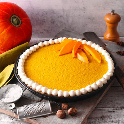 Eine rustikale Kürbis‑Tarte mit Cremehaube liegt auf einer Holzplatte, daneben steht ein Hokkaido‑Kürbis.