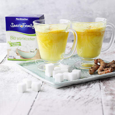 Zwei Glasbecher gefüllt mit leuchtend gelber Goldener Milch stehen auf einem hellen Holzbrett; davor liegen Zuckerwürfel und frische Kurkumawurzeln, im Hintergrund eine Packung Bio-Würfelzucker.