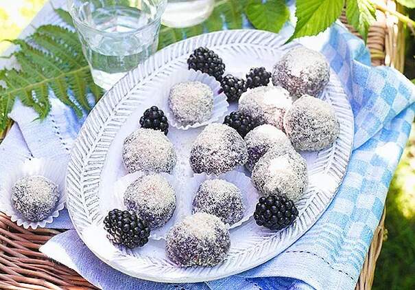 Auf einem weißen, strukturierten Porzellanteller sind mehrere runde Pralinen arrangiert. Die Pralinen haben eine weiße Kokosbedeckung und sind mit frischen, schwarzen Brombeeren kombiniert, die oben auf einigen Pralinen sitzen. 