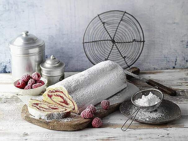 Ein rundes Stollengebäck ist mit Puderzucker bestäubt. Daneben liegen sowohl in einer Schüssel als auch lose frische Himbeeren und ein Sieb mit Puderzucker liegt daneben. 