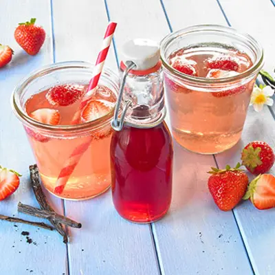 In zwei breiten Gläser, die ebenso gut Einmachgläser sein könntent, ist eine hell-rosane Limonade enthalten, in der Erdbeeren und ein rot gestreifter Strohhalm schwimmen. Davor steht eine kleine Glasflasche mit Sirup drin. 