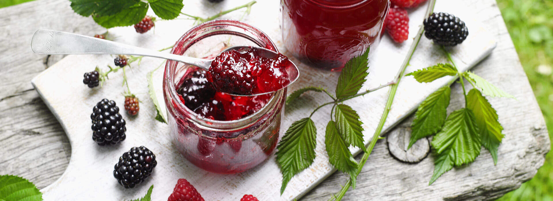 Auf einem weißen Brett steht ein offenes Glas mit tiefrotem Beerengelee, aus dem ein Löffel mit glänzendem Inhalt herausragt. Daneben steht ein weiteres Glas mit Deckel, frische Brombeeren und Himbeeren sowie grüne Blätter sind dekorativ verteilt.