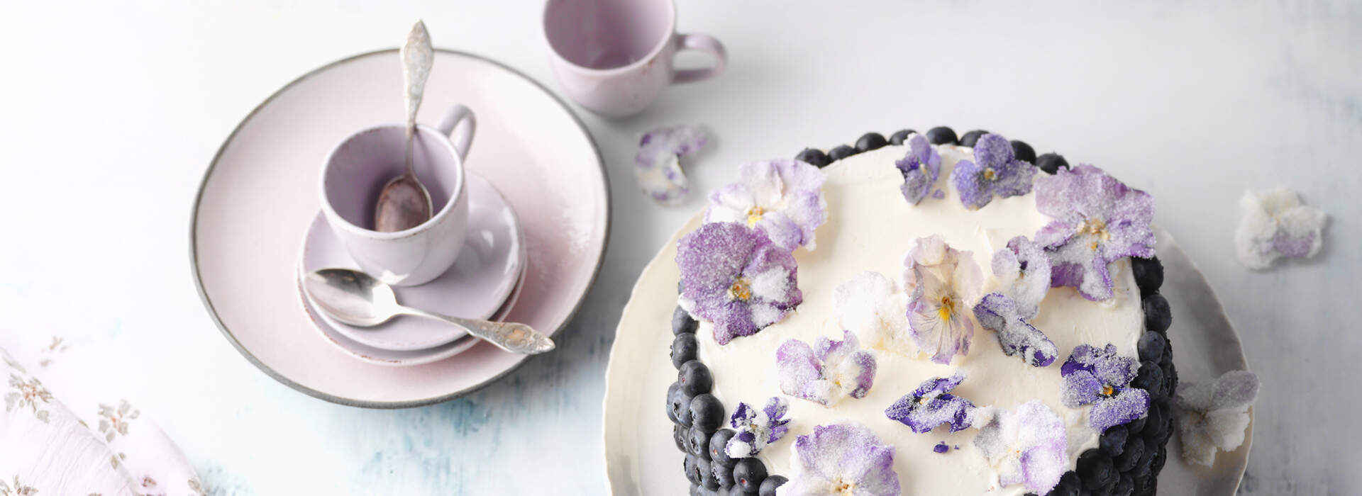 Auf einer hellen Unterlage steht eine runde Torte mit weißer Glasur, deren Rand dicht mit dunklen Heidelbeeren besetzt ist. Die Oberfläche ist mit zarten violetten und weißen kandierten Veilchenblüten übersät, die wie kleine Kunstwerke wirken.