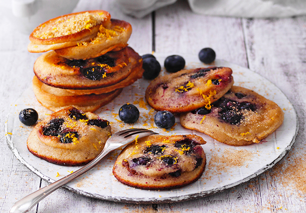 Goldbraune Pancakes sind mit eingebackenen Blaubeeren durchzogen, stapeln sich hoch und sind mit selbstgemachtem Orangenzucker bestreut