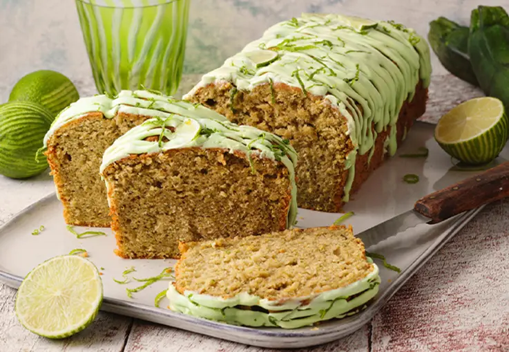 Ein goldbrauner Kasten­kuchen mit geraspelten Zucchini und gemahlenen Mandeln, der mit einem hellgrünen Limettenguss überzogen und mit feinen Limettenzesten bestreut ist