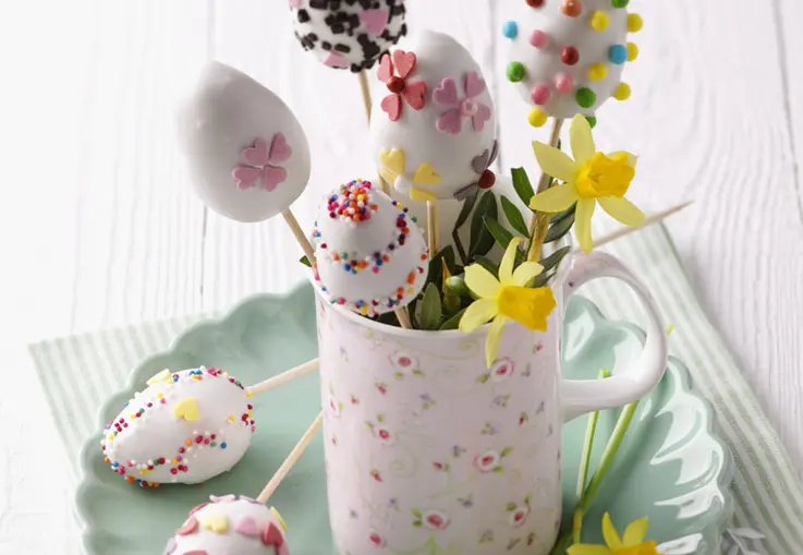 Eierförmige Cake Pops aus Zitronenkuchen, mit weißer Zuckerglasur überzogen und bunt mit Zuckerblüten und Streuseln dekoriert, in einer Tasse arrangiert