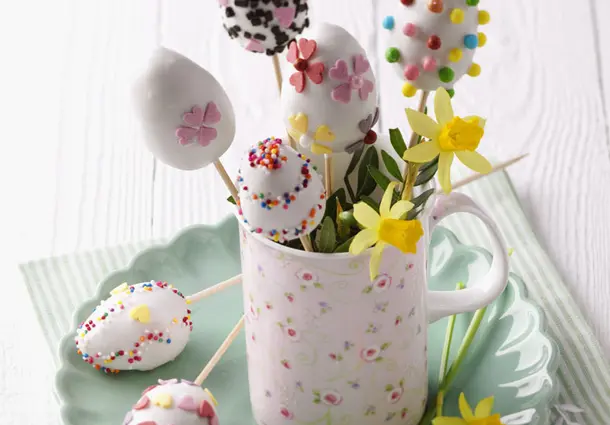 Eierförmige Cake Pops aus Zitronenkuchen, mit weißer Zuckerglasur überzogen und bunt mit Zuckerblüten und Streuseln dekoriert, in einer Tasse arrangiert