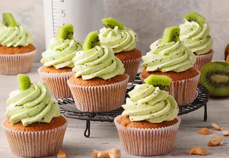 Zarte Cashew‑Cupcakes mit goldenem Teig, getoppt mit einer hellgrünen Kiwi‑Buttercreme und Kiwi-Stückchen, arrangiert auf einem Gitter