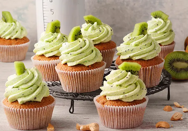 Zarte Cashew‑Cupcakes mit goldenem Teig, getoppt mit einer hellgrünen Kiwi‑Buttercreme und Kiwi-Stückchen, arrangiert auf einem Gitter