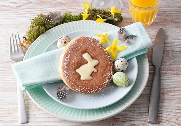 Ein ovaler Osterkeks mit Hasen‑Motiv und Puderzucker bestäubt, liegt auf einem Teller mit bunt gefärbten Eiern und Blüten