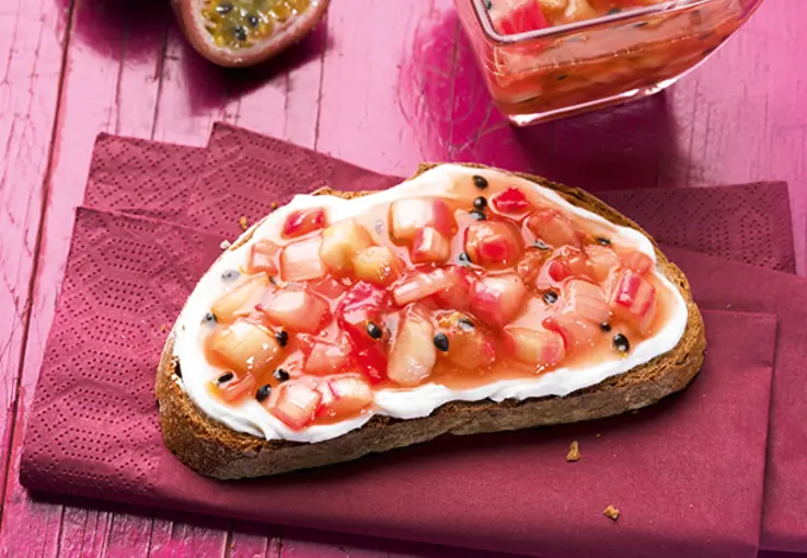Eine Scheibe Brot mit Frischkäse ist mit Rhabarber‑Maracuja‑Konfitüre belegt; daneben liegen halbierte Passionsfrüchte und ein Glas Konfitüre.