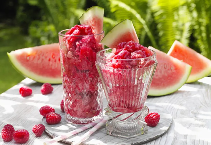 Eisgekühlter rosafarbener Slush aus Wassermelone und Himbeeren im Glas mit frischer Melonenspalte.