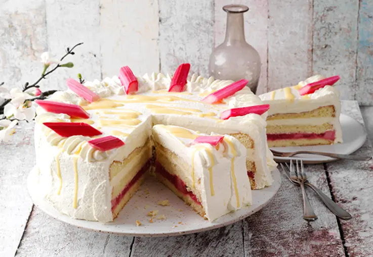 Eine mehrschichtige Rhabarber‑Eierlikör‑Torte aus hellem Biskuit mit Rhabarberkompott und Eierlikörcreme, oben mit Sahne und rosa Rhabarberstücken dekoriert.