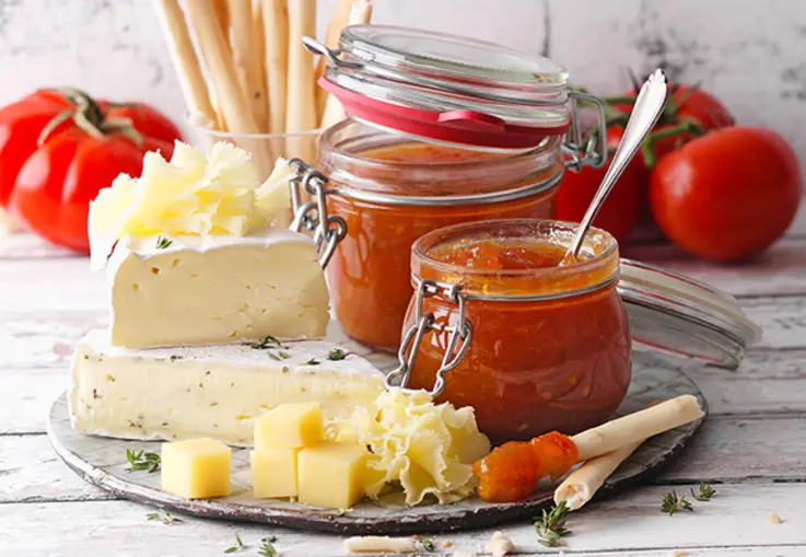 Zwei Gläser Tomaten-Mirabellen-Aufstrich stehen auf einem Brett; daneben Käse, Tomaten, Mirabellen und Brotstangen