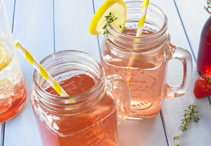 Mason‑Jar‑Gläser mit rosa Erdbeersirup, Zitronenscheiben, Zitronenthymianzweigen und frischen Erdbeeren dekoriert