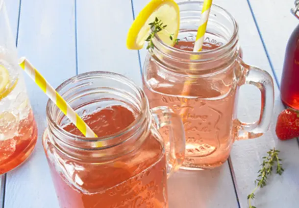 Mason‑Jar‑Gläser mit rosa Erdbeersirup, Zitronenscheiben, Zitronenthymianzweigen und frischen Erdbeeren dekoriert