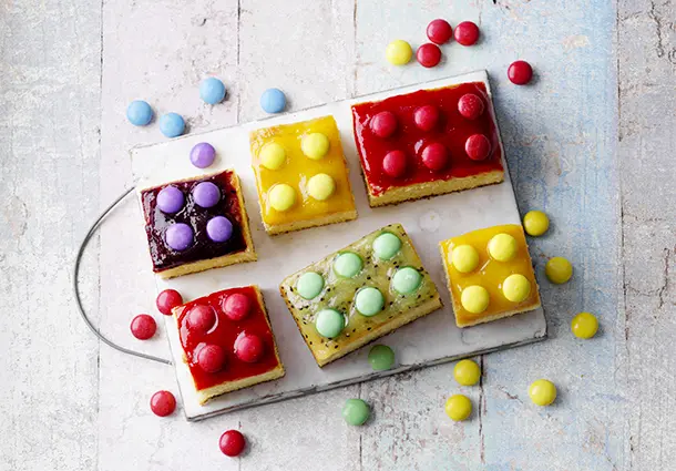 Kleine Kuchenwürfel mit bunten Fruchtglasuren in Rot, Gelb und Violett, dekoriert mit Schokolinsen.