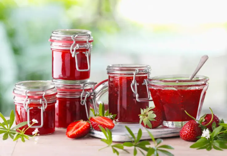 Gläser mit glänzender Erdbeer-Waldmeister-Konfitüre stehen zwischen frischen Erdbeeren und grünen Waldmeisterblättern.