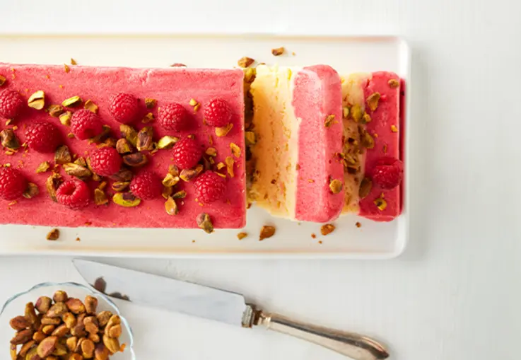Ein längliches Parfait zeigt zwei Schichten: cremiges weißes Schokoladeneis und leuchtendes Himbeersorbet, garniert mit Pistazien und Himbeeren