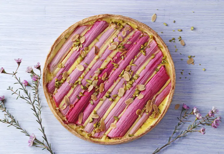 Eine runde Mürbeteig‑Tarte mit Vanille-Schmandcreme, belegt mit länglichen Rhabarberstreifen und mit gehackten Pistazien bestreut