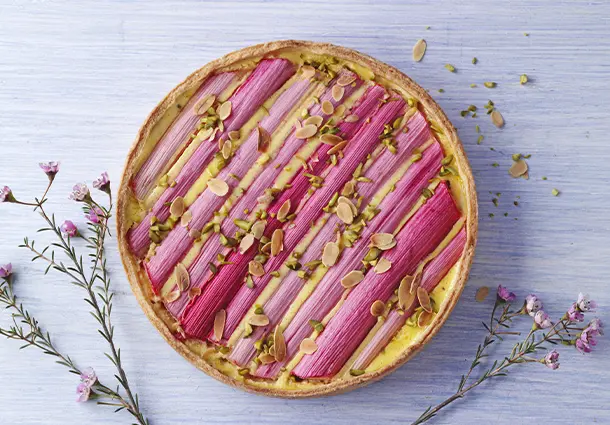 Eine runde Mürbeteig‑Tarte mit Vanille-Schmandcreme, belegt mit länglichen Rhabarberstreifen und mit gehackten Pistazien bestreut