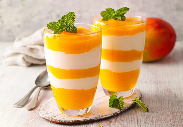 Zweifarbiges Dessert im Glas aus schichtweise gefülltem Mangopüree und cremigem griechischem Joghurt, oben mit frischer Minze dekoriert