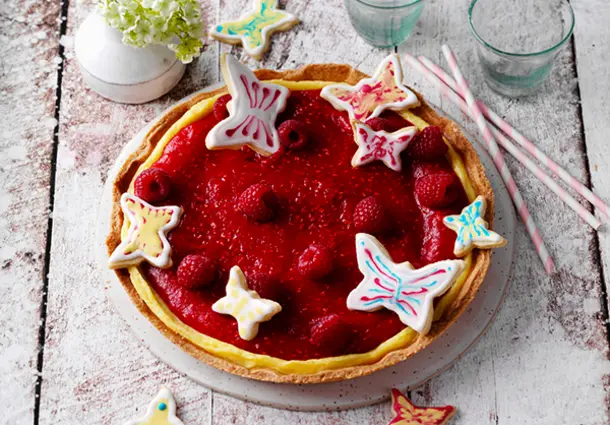 Tarte mit rotem Himbeerspiegel, dekoriert mit Schmetterlingskeksen und frischen Beeren.