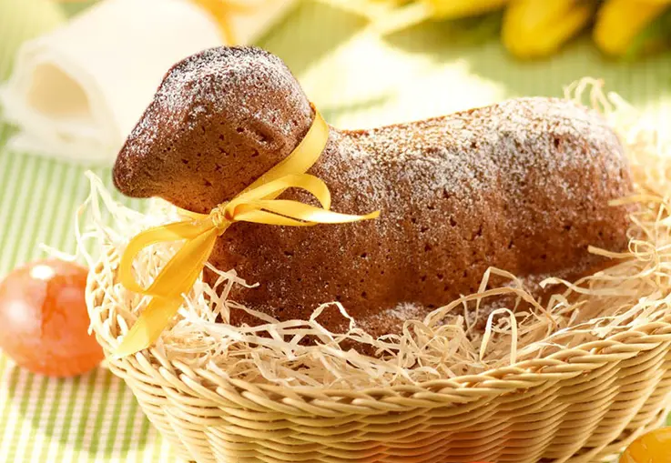 Ein in Lammform gebackener Rührkuchen mit Mandeln, mit Puderzucker bestäubt und mit einer gelben Schleife geschmückt, in einem Korb auf Stroh
