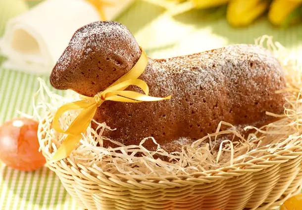 Ein in Lammform gebackener Rührkuchen mit Mandeln, mit Puderzucker bestäubt und mit einer gelben Schleife geschmückt, in einem Korb auf Stroh
