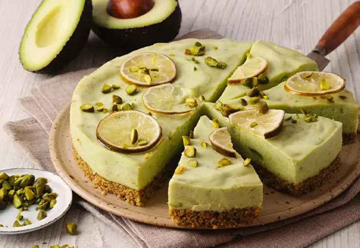 Runder No‑Bake‑Cheesecake mit knusprigem Pistazien-Keksboden, cremiger Avocado-Limetten-Füllung und dekorativen Limettenscheiben sowie gehackten Pistazien