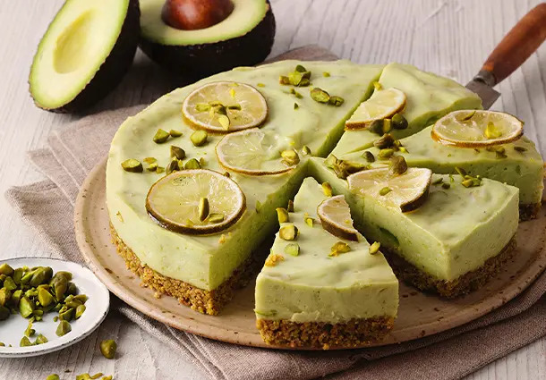Runder No‑Bake‑Cheesecake mit knusprigem Pistazien-Keksboden, cremiger Avocado-Limetten-Füllung und dekorativen Limettenscheiben sowie gehackten Pistazien
