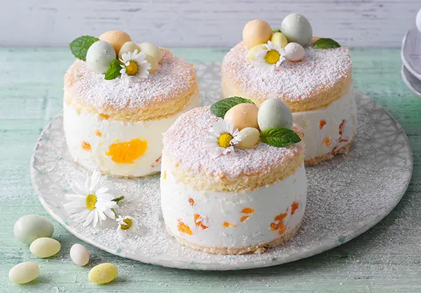 Runde Quarktörtchen mit Biskuitboden und Quark‑Mandarinen‑Creme, mit Puderzucker bestäubt und mit kleinen Zuckereiern und Blüten garniert