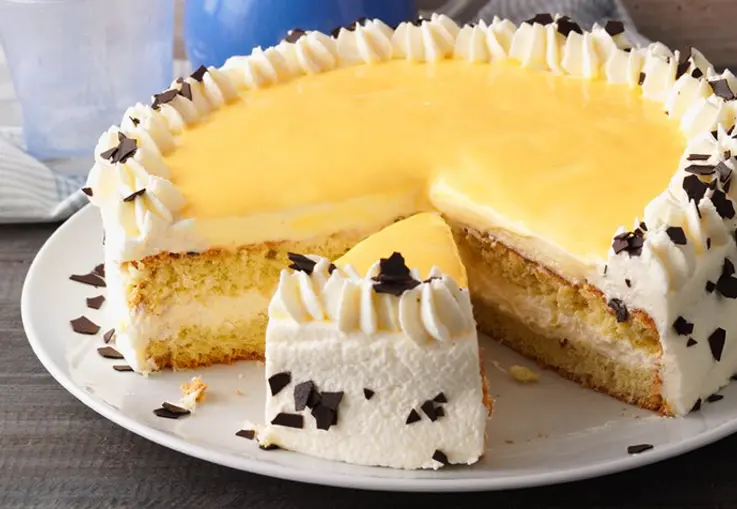Eine runde Torte mit hellen Biskuitböden, Sahnecreme und gelbem Eierlikörguss; die Seiten sind mit Schokoladenraspeln bestreut, oben sitzen Sahnerosetten