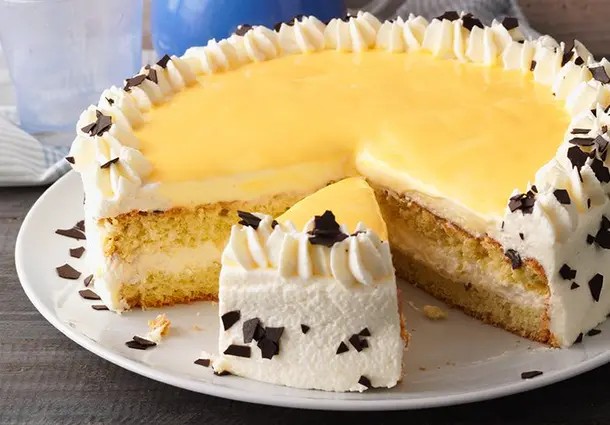 Eine runde Torte mit hellen Biskuitböden, Sahnecreme und gelbem Eierlikörguss; die Seiten sind mit Schokoladenraspeln bestreut, oben sitzen Sahnerosetten