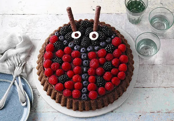 Runde Torte mit heller Creme und roten sowie dunklen Beeren, dekoriert als Marienkäfer mit Schokodetails.