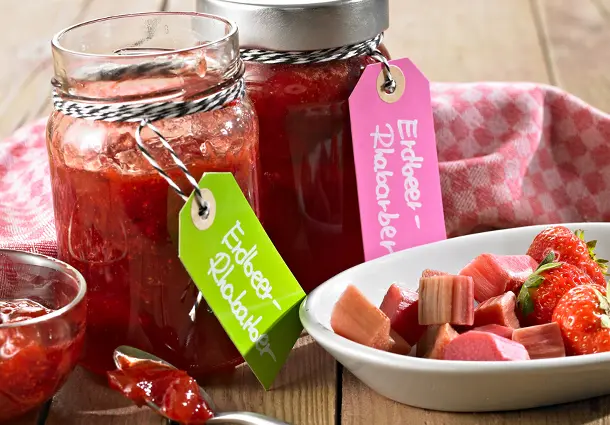 Gläser Erdbeer‑Rhabarber‑Konfitüre mit bunten Etiketten stehen neben einer Schale mit geschnittenen Erdbeeren und Rhabarber.