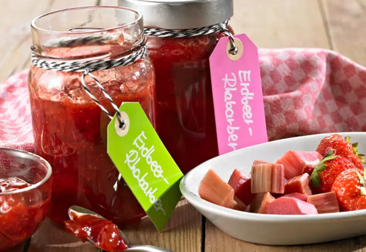Gläser Erdbeer‑Rhabarber‑Konfitüre mit bunten Etiketten stehen neben einer Schale mit geschnittenen Erdbeeren und Rhabarber.