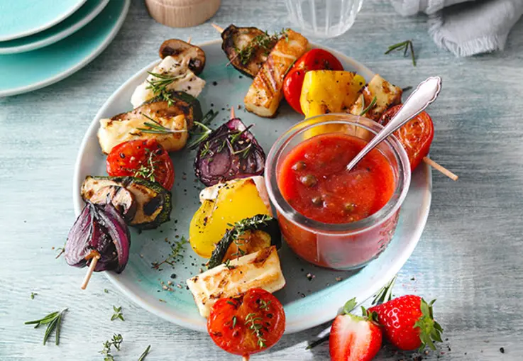 Kleines Glas mit rotem Erdbeer-Tomatenketchup steht neben Gemüsespießen und grünen Pfefferkörnern.