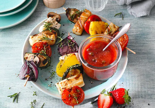 Kleines Glas mit rotem Erdbeer-Tomatenketchup steht neben Gemüsespießen und grünen Pfefferkörnern.