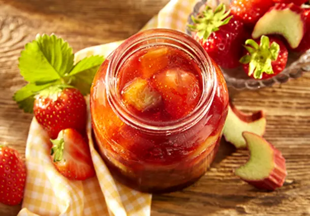 Ein Glas Erdbeer‑Rhabarber‑Konfitüre mit sichtbaren Fruchtstücken steht auf einem Brett, daneben frische Erdbeeren und Rhabarberstücke.