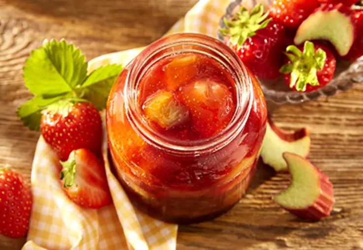 Ein Glas Erdbeer‑Rhabarber‑Konfitüre mit sichtbaren Fruchtstücken steht auf einem Brett, daneben frische Erdbeeren und Rhabarberstücke.