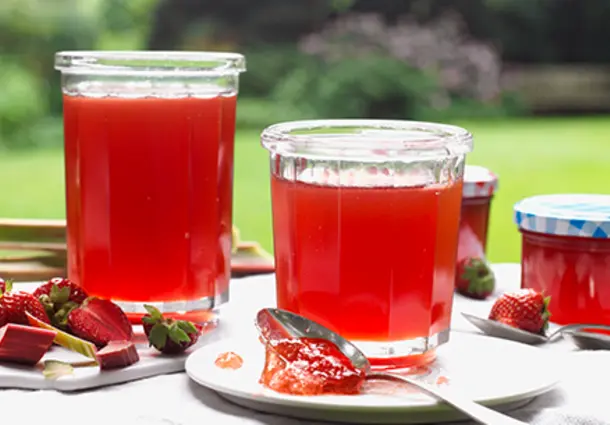 Mehrere Gläser rotes Rhabarber‑Erdbeer‑Gelee stehen auf einem Holzbrett, umgeben von frischen Erdbeeren und Rhabarberstücken.