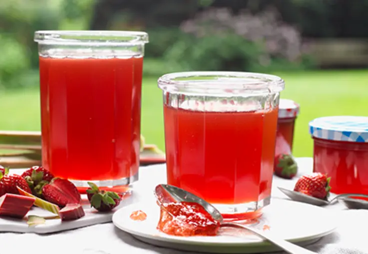 Mehrere Gläser rotes Rhabarber‑Erdbeer‑Gelee stehen auf einem Holzbrett, umgeben von frischen Erdbeeren und Rhabarberstücken.