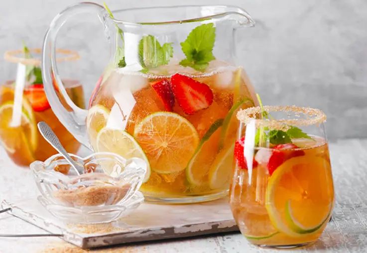 Großer Krug mit Zitronen-Limetten-Eistee, Eiswürfeln und frischen Zitrusscheiben steht auf einem Tisch.